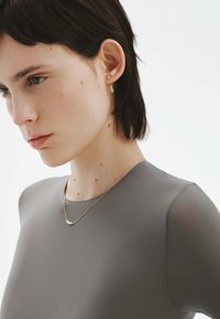 Top gris ajustado con textura suave, escote redondo y mangas transparentes. Lleva un delicado collar de oro y pequeños pendientes de oro.