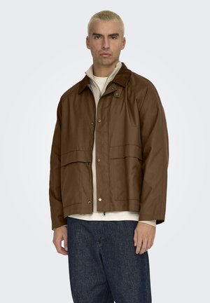 Uomo con capelli biondi corti che indossa una giacca marrone sopra un maglione beige con zip e jeans blu scuro, in piedi davanti a uno sfondo uniforme.