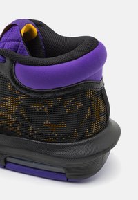 Baskets noires avec des accents violets, fabriquées en tissu en maille et dotées d'une semelle texturée. Des détails jaunes visibles à travers la maille ajoutent un contraste.