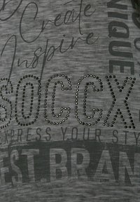 Ciemnoszara koszulka z teksturowanym wzorem. Zawiera wyraźny, duży napis "SOCCX" ozdobiony czarnymi, okrągłymi kryształkami. Dodatkowy tekst wydrukowany w różnych czcionkach.