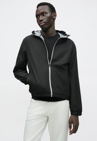 Μαύρο μπουφάν με κουκούλα από ελαφρύ υλικό, με εσωτερική επένδυση σε ασημί χρώμα, φερμουάρ μπροστά και ελαστικές μανσέτες και στρίφωμα.