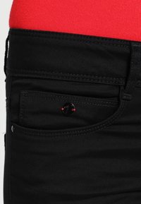 Jeans de mezclilla negros con costuras rojas, con un pequeño bolsillo que tiene un acento de botón negro circular y un acabado de tela ligeramente texturizado.