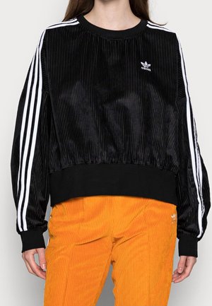 Persona che indossa una felpa Adidas nera in velluto con strisce bianche e pantaloni in velluto a coste arancioni, in piedi davanti a uno sfondo semplice.