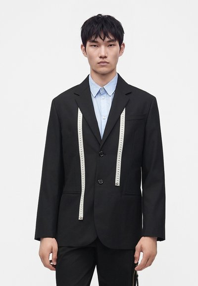 Jeune homme portant un costume noir et une chemise bleu clair avec un ruban à mesurer blanc drapé sur ses épaules, sur un fond uni.