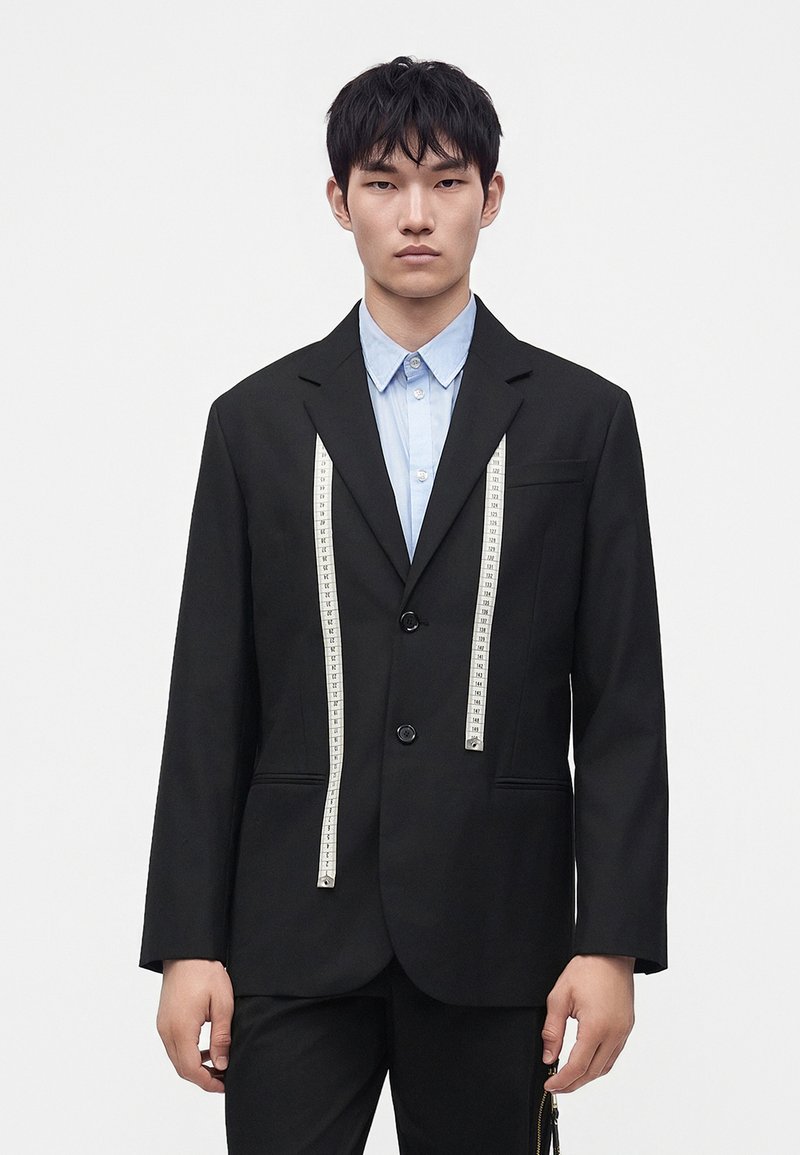 Jeune homme portant un costume noir et une chemise bleu clair avec un ruban à mesurer blanc drapé sur ses épaules, sur un fond uni.