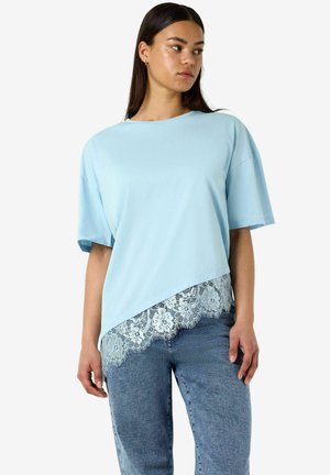 Vrouw draagt lichtblauw asymmetrisch T-shirt met kanten zoom en blauwe jeans, staand tegen een effen witte achtergrond.