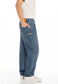 Light blue denim jeans with a relaxed fit, featuring a back pocket, orange brand label, and cuffed hems, paired with white sneakers.