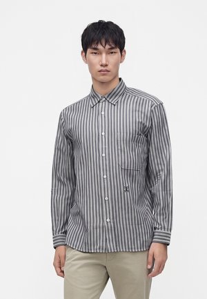 Jeune homme portant une chemise à rayures verticales grises et blanches avec une poche sur le devant et un pantalon beige, debout devant un fond uni.