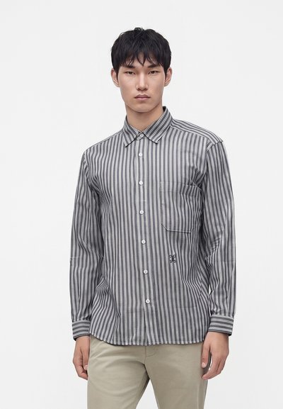 Jeune homme portant une chemise à rayures verticales grises et blanches avec une poche sur le devant et un pantalon beige, debout devant un fond uni.