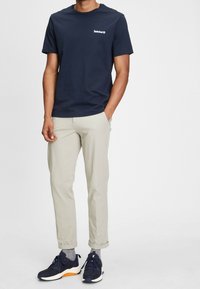 Navybomull t-shirt med Timberland-logotyp, ljusbeige byxor och mörka sneakers med orange detaljer. Slät textur, vanlig passform.