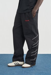 Άτομο που φοράει μαύρο παντελόνι Adidas με κόκκινο λογότυπο και μπεζ αθλητικά παπούτσια Adidas με λευκές ρίγες, στέκεται σε μπλε χαλί.