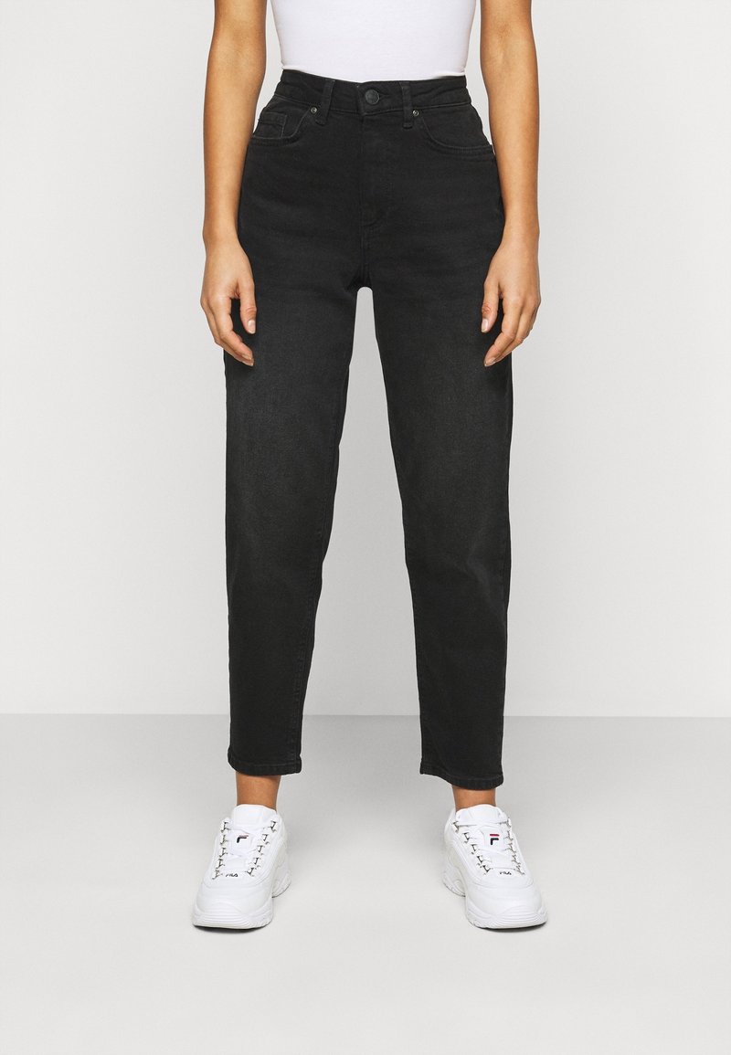 Persona que lleva puestos unos pantalones negros de cintura alta y unas deportivas blancas de FILA, de pie contra un fondo claro y sencillo.