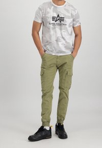 Koszulka z krótkim rękawem w kolorze jasnoszarym z nadrukiem w kamuflaż, z czarnym logo; oliwkowe spodnie cargo z bocznymi kieszeniami; czarne sneakersy z białymi akcentami.