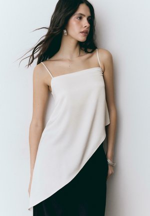 Frau mit langen dunklen Haaren trägt ein weißes asymmetrisches ärmelloses Oberteil und einen schwarzen Rock, sie steht vor einem schlichten hellen Hintergrund.