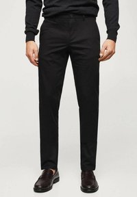 Mann trägt schwarze Slim-Fit-Hose, dunkelbraune Ledermokassins und einen dunklen Langarmpullover, steht vor einem schlichten Hintergrund.