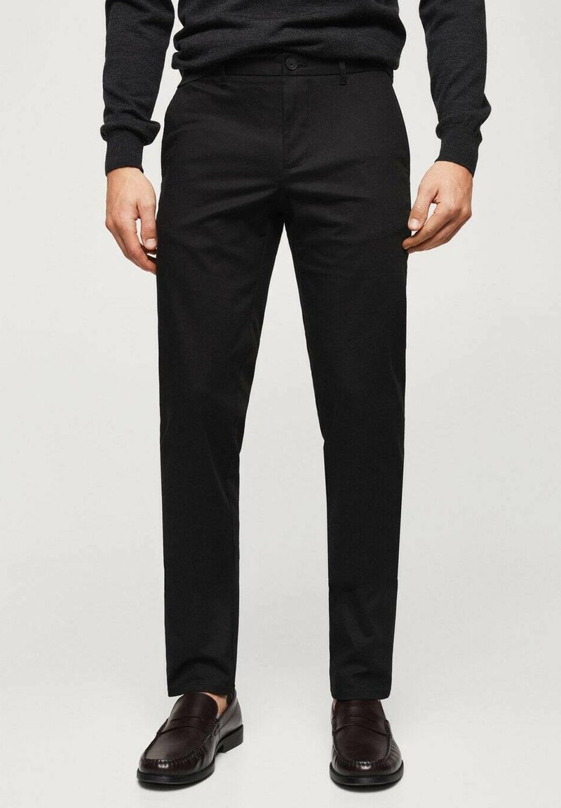 Mann trägt schwarze Slim-Fit-Hose, dunkelbraune Ledermokassins und einen dunklen Langarmpullover, steht vor einem schlichten Hintergrund.