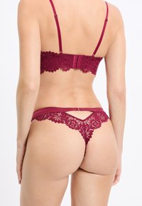 Bordeauxrotes Spitzen-Dessous-Set mit einem Bralette mit gewellten Kanten und passendem Tanga mit floralen Mustern, das eine zarte Textur zeigt.