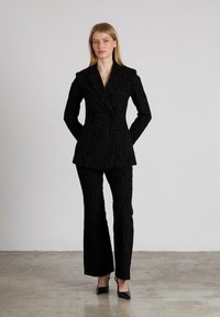 Traje negro a medida con un patrón floral texturizado, que presenta un blazer de doble botonadura y pantalones acampanados, complementados con tacones negros puntiagudos.
