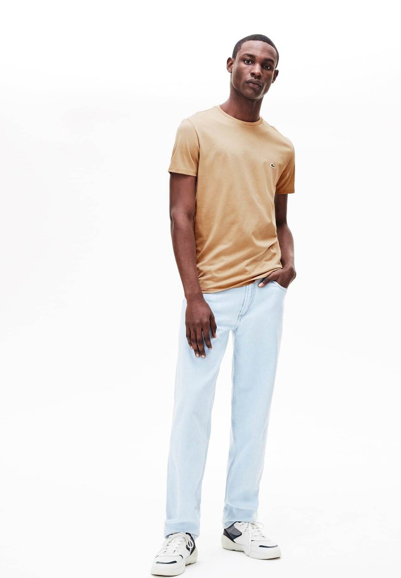 T-shirt a maniche corte color marrone chiaro con logo, abbinata a jeans dritti blu chiaro e sneakers bianco e nero su uno sfondo neutro.