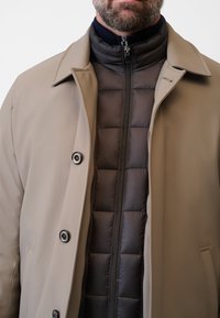 Beige ytterkappa med en slät textur och stora knappar, lagd över en svart vadderad innerjacka med dragkedja.