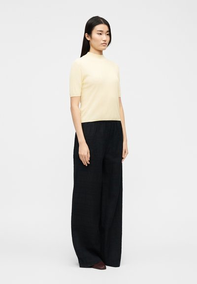 Γυναίκα που στέκεται με μπεζ κοντομάνικο μπλουζάκι με ψεύτικο λαιμόκοψη, μαύρο παντελόνι με φαρδιά πόδια και σκούρα καφέ μυτερά παπούτσια μπροστά από απλό φόντο.