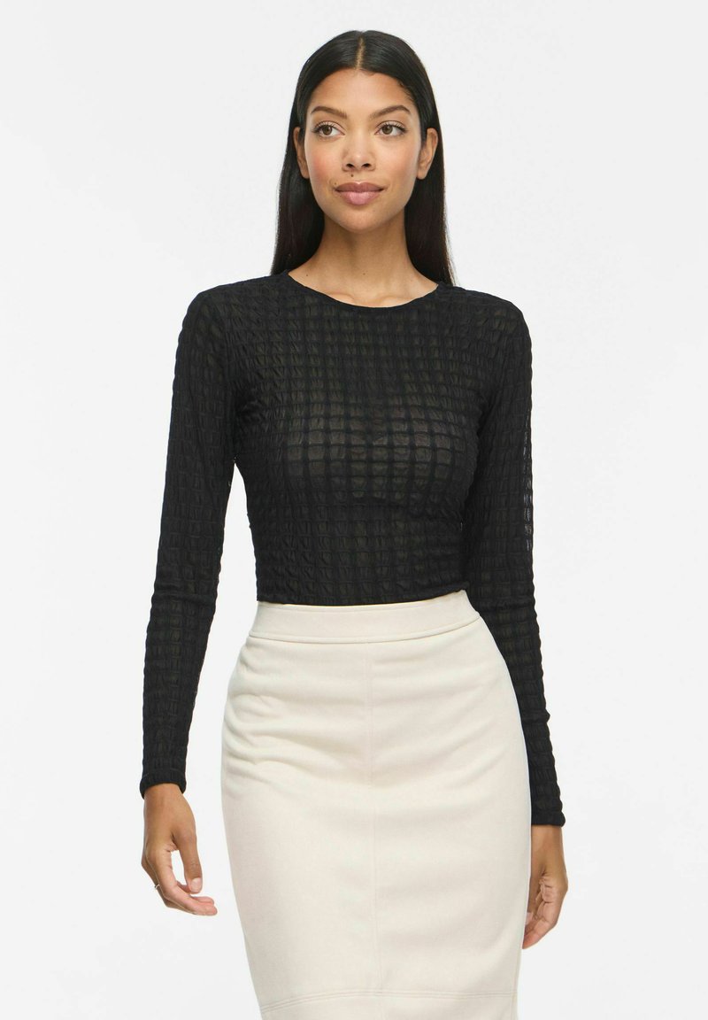 Femme aux longs cheveux foncés portant un haut noir texturé à manches longues et une jupe crayon crème taille haute, debout devant un fond blanc uni.