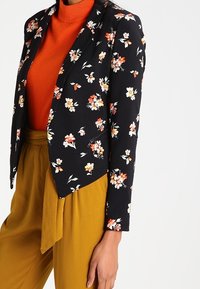 Svart blommig kavaj med oranga och vita blommor, lagd över en orange polotröja och senapsgula byxor, med en figurnära passform.