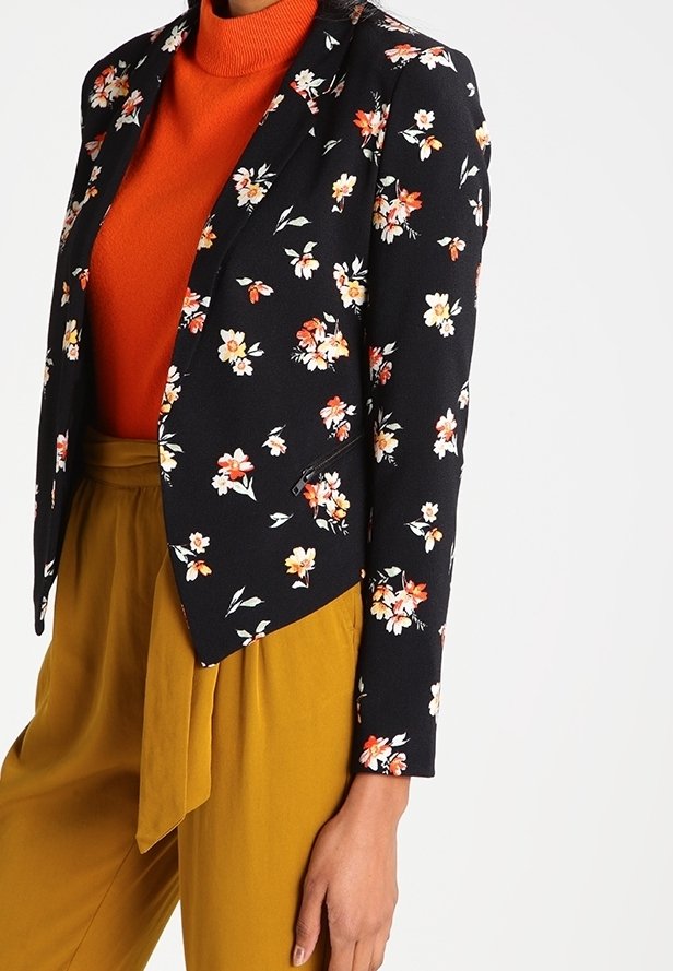 Svart blommig kavaj med oranga och vita blommor, lagd över en orange polotröja och senapsgula byxor, med en figurnära passform.
