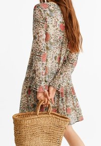 Kleid mit floralem Muster, langen Ärmeln, taillierter Taille und volantiertem Saum. Das Model hält eine geflochtene Korbtasche. Die Farben umfassen Grün, Pink und Beige.