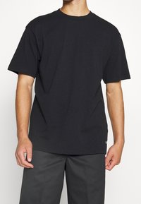 T-shirt nera in cotone con maniche corte, scollo rotondo, vestibilità rilassata e un piccolo logo sul fondo. Abbinato a pantaloni grigi.