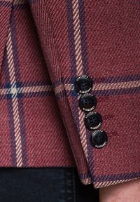 Wełniana marynarka w kolorze burgundowym w kratkę, z czterema czarnymi guzikami z białym napisem. Ciekawe kontrastowe przeszycia w kolorze niebieskim i białym.