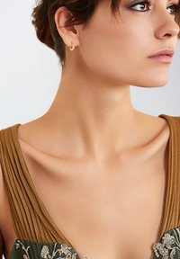 Gouden hoepeloorbel, geribbeld bruin stoffen schouderbanden, groene geborduurde stof met bloemmotieven, soepele huidtextuur, neutrale achtergrond.