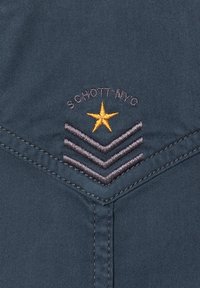 Donkerblauwe stof met een chevrondesign, voorzien van geborduurde strepen in grijs en een ster in goud, samen met de tekst "SCHOTT NYC" in donkere draad.