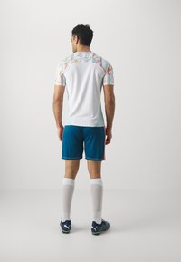 Camiseta deportiva con un patrón geométrico colorido en los hombros, cuerpo blanco, combinada con pantalones cortos en azul verdoso, calcetas blancas y zapatillas deportivas oscuras.