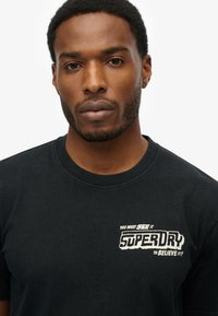 T-shirt in cotone nero con scollatura rotonda e una grafica frontale in testo bianco che recita "DEVI VEDERLO SUPERDRY PER CREDERCI!"