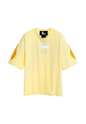 Camiseta de manga corta amarillo pálido con diseños de plumas naranjas en ambas mangas y una pequeña etiqueta central debajo del cuello.
