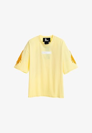 Camiseta de manga corta amarillo pálido con diseños de plumas naranjas en ambas mangas y una pequeña etiqueta central debajo del cuello.