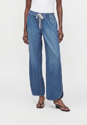 Γυναίκα που φοράει φαρδιά μπλε τζιν παντελόνια με ελαστική μέση και λευκό κορδόνι, συνδυασμένα με λευκό τοπ και μαύρα πλατφόρμα σανδάλια.