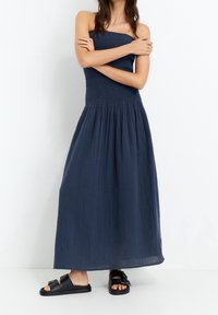 Robe longue bleu marine avec un corsage froncé et une jupe fluide. Fabriquée en tissu léger. Associée à des sandales plates noires avec une boucle.