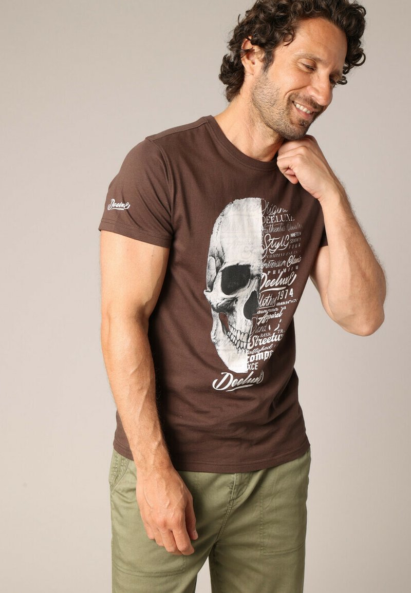 T-shirt en coton marron avec un grand motif de skull blanc accompagné d'éléments textuels. Le t-shirt a des manches courtes et un designcol rond.