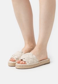 Sandales slip-on tissées beiges avec un dessus en tissu tressé, semelle en jute et design à bout ouvert, présentant une finition texturée et naturelle.