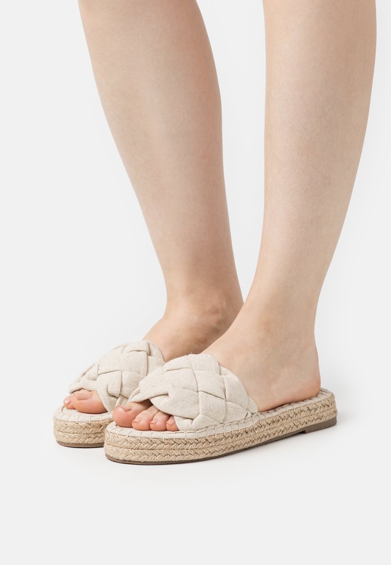 Sandales slip-on tissées beiges avec un dessus en tissu tressé, semelle en jute et design à bout ouvert, présentant une finition texturée et naturelle.