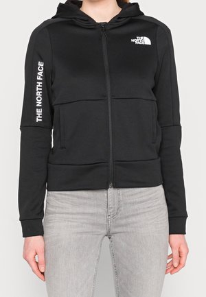 Czarna bluza z kapturem na zamek z logo The North Face na piersi i rękawie, noszona z jasnoszarymi dżinsami, ręce swobodnie opuszczone wzdłuż ciała.