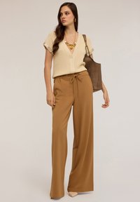 Beige pletený top s výstřihem do V, kombinovaný se širokými, vysokými hnědými kalhotami s tkaničkou. Doplněno je chunky náhrdelníkem a kabelkou.
