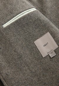 Brown and cream herringbone fabric with a white piped pocket. Label: "NEXT," "REGULAR FIT," with care instructions and size details.