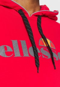 Sweat à capuche rouge avec cordons noirs et blancs, arborant un grand texte gris "ellesse" et un logo demi-cercle orange sur la poitrine.