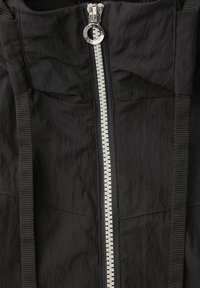 Sort jakke set forfra med sølvlynlås og sorte snore på en tekstureret stofbaggrund.