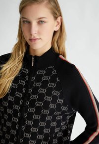Giacca nera con zip caratterizzata da un motivo logo ripetuto, materiale a maglia testurizzato e accenti striati rossi e dorati contrastanti sulle maniche.