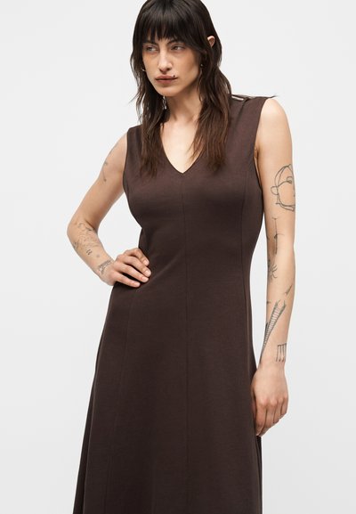 Γυναίκα με μακριά σκούρα μαλλιά που φοράει ένα αμάνικο καφέ φόρεμα, στέκεται με το ένα χέρι στη μέση, δείχνοντας τατουάζ στο χέρι.