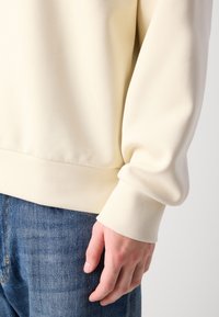 Main droite reposant à côté du torse, vêtue d'un sweat-shirt crème à manches longues et d'un jean en denim bleu, sur un fond uni.